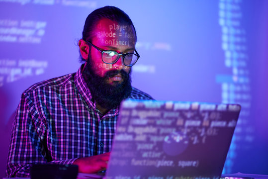 Serious Bearded IT Man In Eyeglasses Sitting At The Table And Using Laptop Computer For Developing New Website In Dark Office