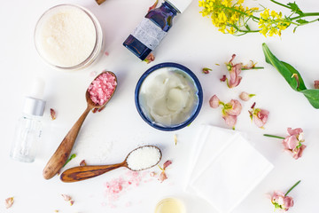Solid shea butter in a black jar with a wooden spoon on a white background, next to white and pink petals, green plant twigs. Copy space.Top view 