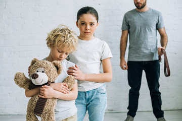 partial view of abusive father with belt and sad kids with teddy bear