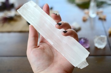 Large Selenite Rod, Holding Crystal over wooden table. Women's hand holding selenite in natural lighting. Healing selenite chunk, large witch wand. Wiccan healing crystal tools, zen meditation stones.