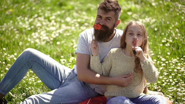 Family spend leisure outdoors. Dad and daughter sits on grass at grassplot, green background. Child and father posing with pipe and lips photo booth attributes. Fatherhood concept.