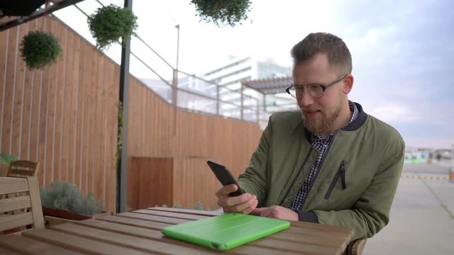 Adult man is resting in open terrace of cafe, calling by video in smartphone