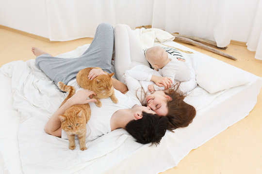 portrait of happy parent playing with their kid. Warm and cozy scene in real life interior. Lifestyle photo. Mom,dad, son and their domestic ginger cat lying on white bed