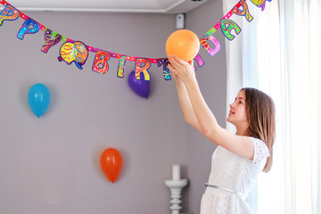 Happy preteen girl hanging up balloons decorating house preparing to birthday party. Happy birthday string.