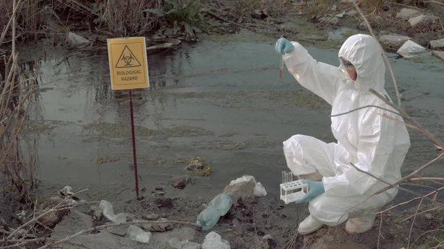 bio-hazard in nature, hazmat chemist into protective suit and mask taking infected water sample in test tubes for testing in contaminated river with pointer sign biological hazard