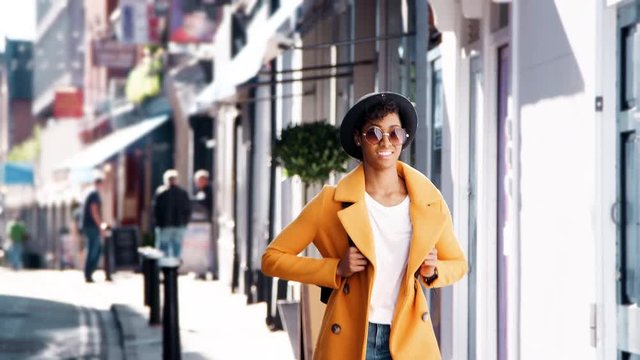 Fashionable young black woman wearing blue jeans and an unbuttoned yellow pea coat walking on a street past shops on a sunny day, smiling, close up