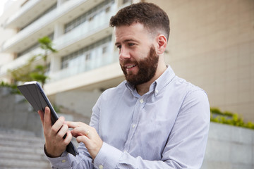 Bearded young man standing in the city and working online, he looking on digital tablet and smiling