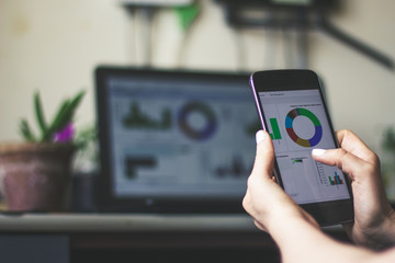 Young woman hand holding a smartphone with a chart on the screen with a laptop in background – Mobile phone used for statistics and annualizing graphics