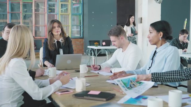 The business team holds a meet up at a large table in the office. One of the staff members gets up and leaves. Creative office interior. Coworking startup team. Office workers