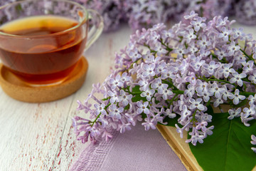 Obraz na płótnie Canvas Spring flowers of lilac and leaves , a mug of tea and a book on a wooden background. Decorative frame, close-up, top view.