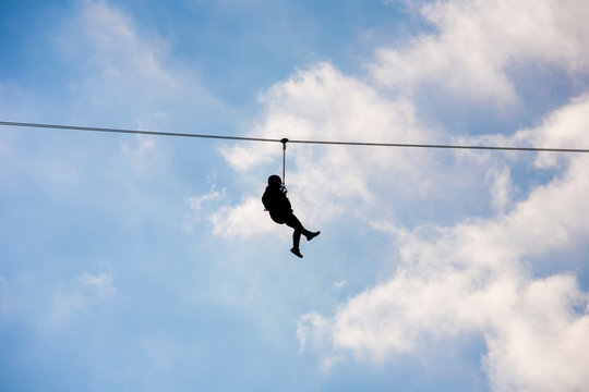 Silhouette of a hanging man on a zip line in front of a cloudy blue sky background