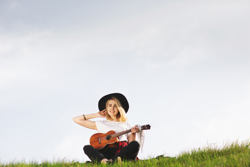 Outdoor portrait of a young beautiful woman in black hat, playing guitar. Space for text