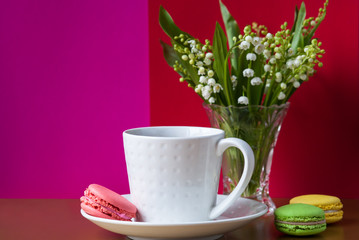 French multicolored cakes macarons and a cup of tea. A bouquet of lily of the valley in a crystal vase.