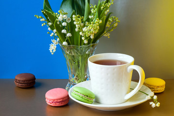 French multicolored cakes macarons and a cup of tea. A bouquet of lily of the valley in a crystal vase.