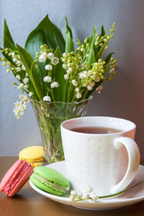 French multicolored cakes macarons and a cup of tea. A bouquet of lily of the valley in a crystal vase.
