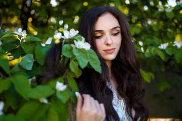 a teenager girl with dark long wavy hair in white dress standing sad in the apple garden