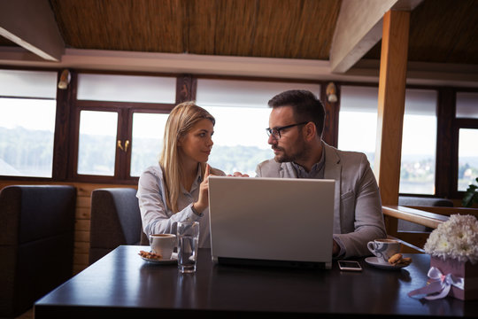 A handsome businessman and beautiful blond businesswoman talking while sitting in a restaurant in front of a laptop and drinking coffee.