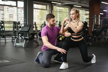 Zelfklevend Fotobehang Sporty girl doing squats exercises with assistance of her personal trainer at public gym. Coaching assistance training concept © ANR Production