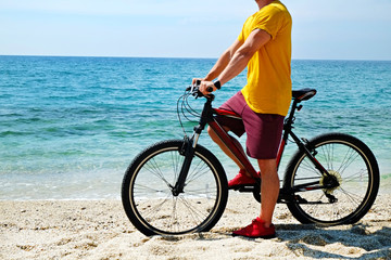 Fototapeta premium Young man with fit body riding the mtb mountain bike on sandy beach with beautiful azure water sea view. Muscle male wearing bright yellow t-shirt cycling on ocean shore. Close up, copy space.