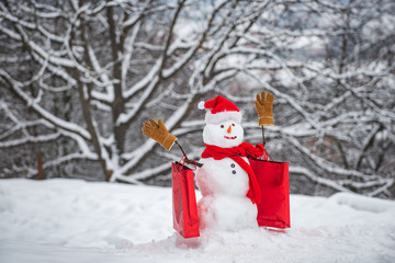 Christmas holidays discounts and winter sale. Snowman in a scarf and hat with shopping bag. Happy snowman with gift boxes standing in winter Christmas landscape.