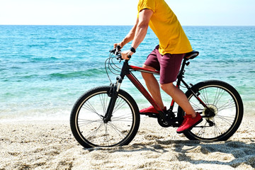 Fototapeta na wymiar Young man with fit body riding the mtb mountain bike on sandy beach with beautiful azure water sea view. Muscle male wearing bright yellow t-shirt cycling on ocean shore. Close up, copy space.