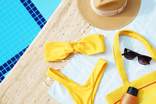 Swimming pool accessories concept. Top view of beach items on deck, bright yellow bikini bathing suit, hipster straw hat, blue mat & sunscreen skincare cream. Colorful beach wear. Flat lay, copy space