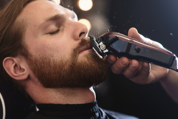 Close-up side view portrait of handsome young bearded caucasian man getting beard grooming in modern barbershop. Hairdresser at work with shaver