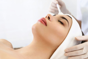 Model in white headband bandage cap lying on couch with closed eyes. Hand of beautician doctor in white glove touching her face with brush. Head and shoulders, healthcare, cosmetology, beauty clinic