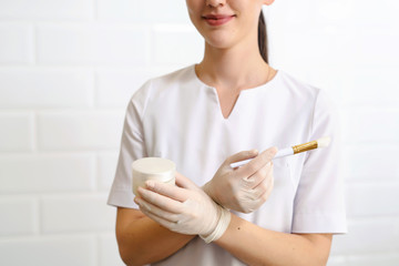 Portrait of young attractive smiling beautician doctor with brush and cream mask pot in hand. Cosmetologist holding medical tools. Beauty clinic or hospital. White background isolated
