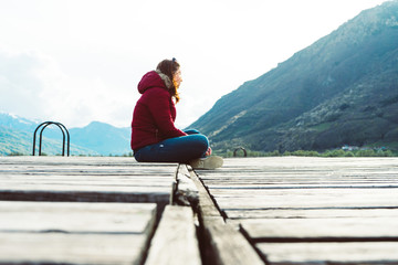 A young girl sits on the edge of a wooden pier and looks into the distance surrounded by mountains in Plav lake