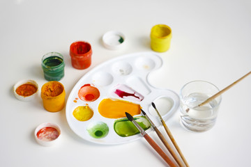 Open jars with gouache of different colors: yellow, orange, red, green, as well as brushes, plastic palette and a glass of water for washing brushes.