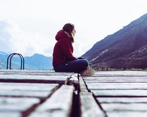 A young girl sits on the edge of a wooden pier and looks into the distance surrounded by mountains...