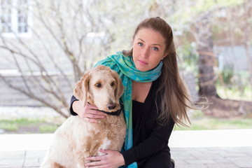 Medium horizontal portrait of pretty blonde windswept young woman wearing a turquoise shawl and with her arm around her golden doodle with soft focus garden in the background