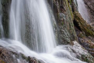 Close up, long exposure view of a sunlit waterfall with soft streams of water falling down red rocks covered by green moss