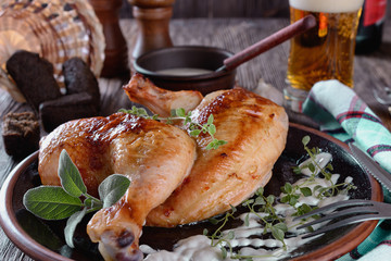 Half  of baked chicken with crispy crust and spicy herbs on a ceramic plate on a wooden table next to small rye-bread buns, white sauce and glass of beer