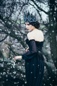 grim lady with pale skin and dark hair beside flowering tree, witch turns with black crow with long luxurious dress and open back and shoulders, gothic image and makeup, cold metal crown and jewels