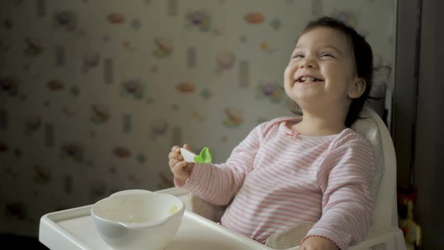 Cute baby gil sitting in feeding chair and eating healthy food on kitchen