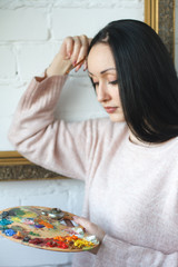 Young woman with long black hair holds a brush and a palette with multi-colored acrylic paints on the background of an empty vintage frame