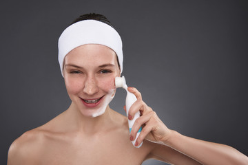 Joyful young woman in white headband using pore cleansing brush after bath