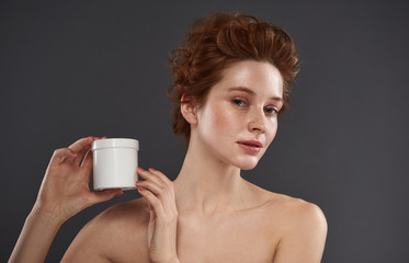 Charming red-haired girl holding white jar of cosmetic cream