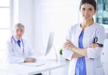 A doctor smiling at the camera with her male colleage in the back of the consulting room in hospital