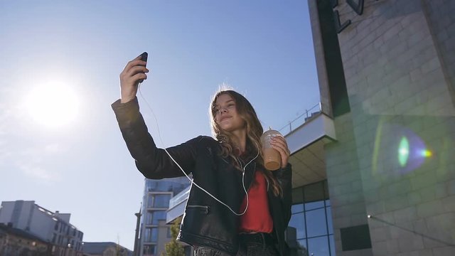 Portrait of the pretty young blonde girl taking selfie photos on the smartphone device camera while walking urban street on sunset. Outdoors