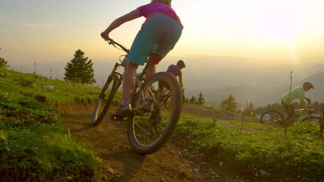 SLOW MOTION, SUN FLARE: Golden sunbeams shine on tourists riding their bicycles along a steep dirt trail. Four mountain bikers ride into a hairpin turn of a mountain path on sunny spring evening.