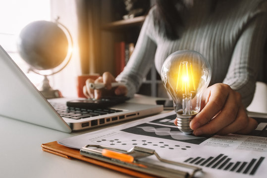 business hand holding lightbulb with using laptop computer and money stack in office. idea saving energy and accounting finance concept  in morning light