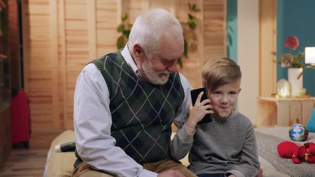 Old carrying grandfather looking for little boy in gray sweater. Nice child holding smartphone in hands and calling mom, laughing, listening to voice, speaking. Inside.