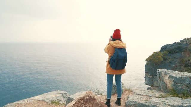 Tourist girl with a backpack standing on the edge of a cliff and shooting video of the sea on an autumn cloudy day. Slow motion.