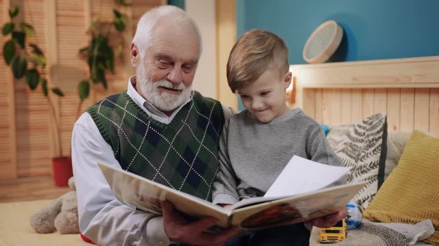 Cute little boy and old gray-haired grandfather viewing pictures in colored book, flipping pages, reading. Good mood, smile. Leisure, education.