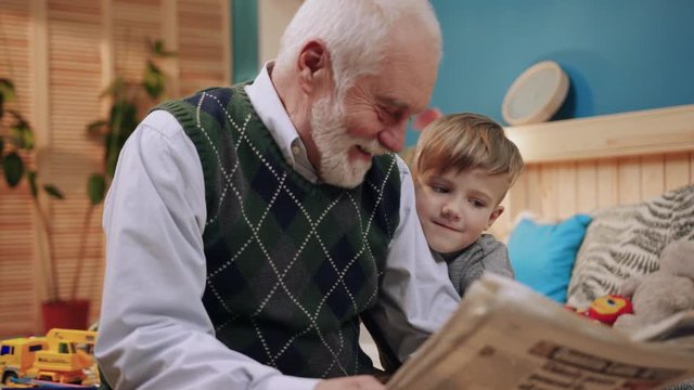 Nice child holding on to hand of grandpa and men reading newspaper together. Close-up footage from below. Indoors. Bedroom. Lifestyle. Love, daily, concept.