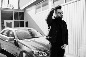 Fashion Arab man wear on black jeans jacket and sunglasses posed against business modern car. Stylish, succesful and fashionable arabian model guy.