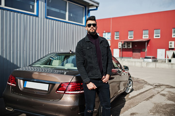 Fashion Arab man wear on black jeans jacket and sunglasses posed against business modern car. Stylish, succesful and fashionable arabian model guy.